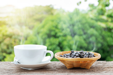 Hot coffee cup and coffee beans in the bamboo plate on the old wooden floor with colorful green nature background.