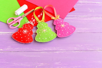 Green, red and pink Christmas trees crafts, scissors, white thread, needle, felt sheets set on a lilac wooden background. Bright Christmas toys sewn from felt and decorated with beads and snowflakes