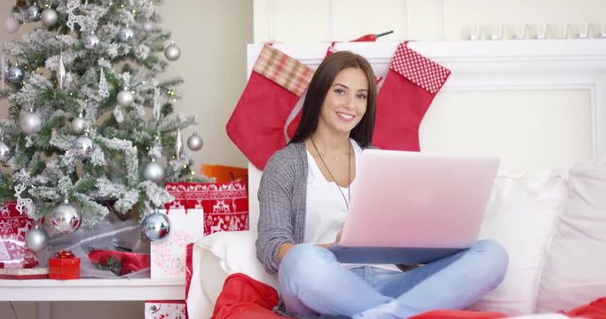 Contented smiling young woman working on her laptop while relaxing over the Christmas holiday in her festive living room at home