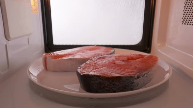 Using microwave oven to defrost salmon steaks before cooking it. Two fresh frozen fish steak on a plate spinning inside the microwave. Version without external lighting for more natural look.