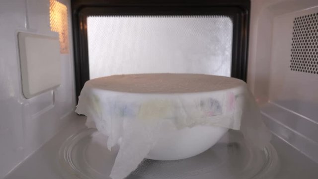 Ceramic bowl with some food covered with a wet towel heating up in the microwave. Dish spinning on turntable tray inside oven.