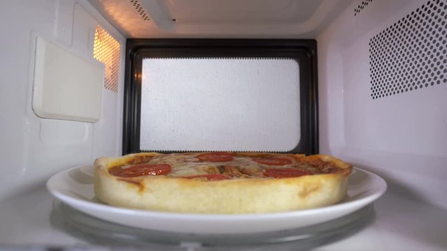 Girl in the kitchen using microwave to reheat baked pizza. She looks inside the oven and takes out plate with heated food.