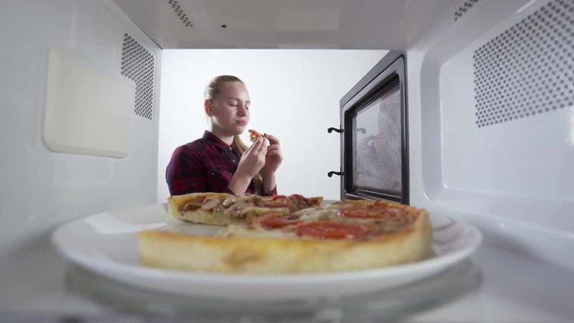Young girl eats a slice of pizza just reheated in the microwave. View through the oven.