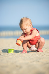 Baby playing on the sandy beach near the sea. Cute little girl in red dress with sand on tropical beach. Ocean coast.