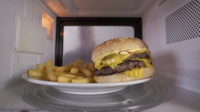  Reheating hamburger with french fries in the microwave. Young girl opens the door and takes out plate with heated lunch. View through the oven.