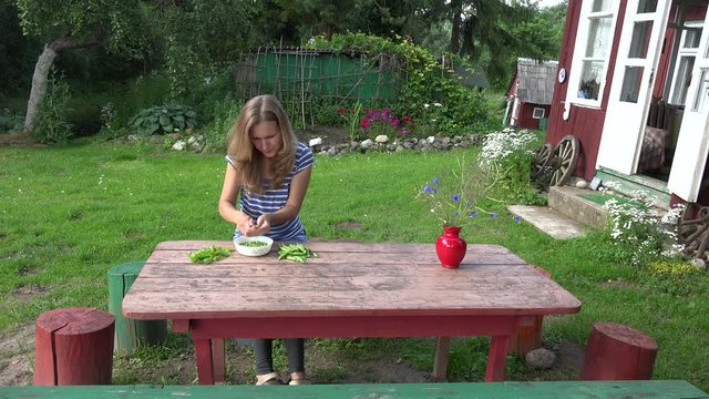 Villager blond girl pod green peas on wooden table near rural house. 4K