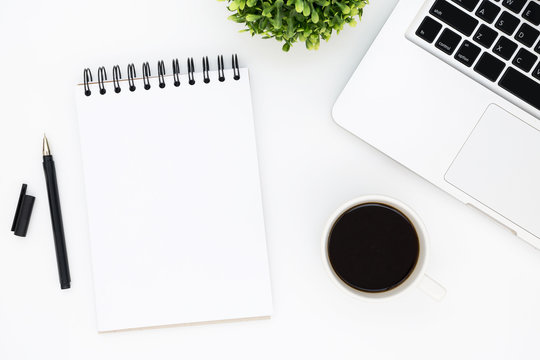 White office desk table with blank notebook page with pen, laptop and coffee. Top view with copy space, flat lay.