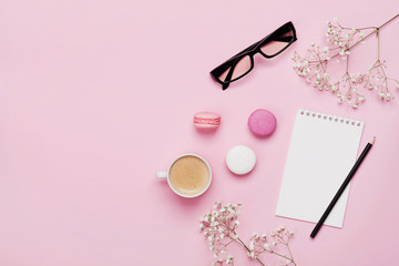 Coffee, cake macaron, clean notebook, eyeglasses and flower on pink table from above. Female working desk. Cozy breakfast. Flat lay style.