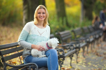 Young woman is sitting on a bench while walking her dog in a park. She is smiling and looking at camera.