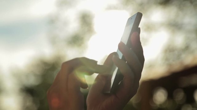 Silhouette of woman hands with smart phone. Typing and touching. Close up shot of mobile cell phone, outdoors in the garden, in nature. Sunset light, sun lens flare, golden hour