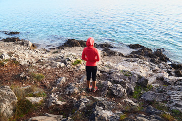 Young fitness woman runner wearing orange jacket and black pants stand on stony beach, relaxing after running and working out outdoors near sea. Girl looking and enjoying view of the water.
