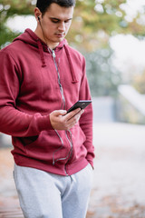 Fototapeta premium Autumn outdoors. Young serious handsome guy with earphones standing with bicycle in park, holding cell phone.