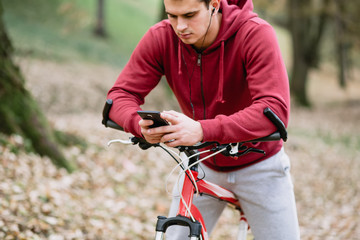 Autumn outdoors. Young serious handsome guy with earphones standing with bicycle in park, holding cell phone.