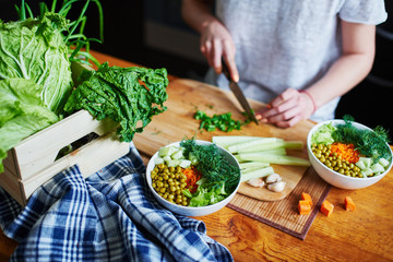 Bowl with green peas, cucumbers, carrots, lettuce and dill standing on a table on a background of hand chopping green onions with knife on wooden Board