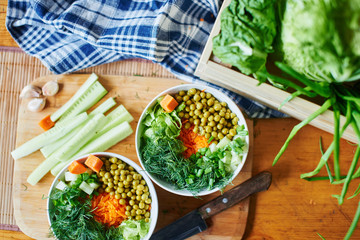 Top view of vegetable mix in bowls with green peas, cucumbers, carrots, lettuce and dill, standing on a wooden table