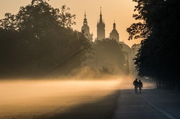 Fototapeta Unidentified men silhouettes in the mist, Blonia meadow in Krakow, Poland, with St Mary's church and Town Hall towers in the background. obraz
