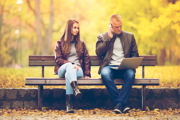Young couple sitting ona a bench in a park on a beautiful Autumn day. Young man is working on his...