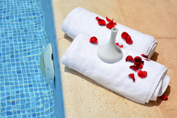 Rose petals and a decorative vase on a white towel by the pool 