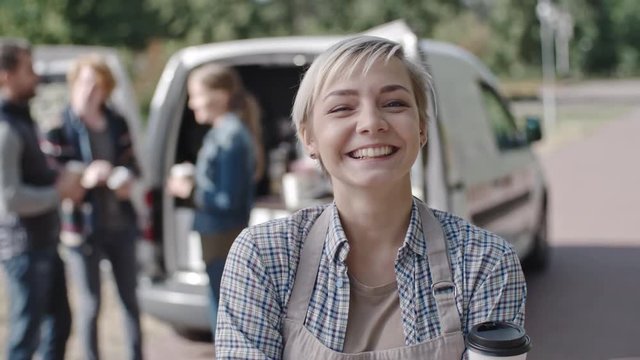 Portrait of blond female owner of mobile coffee van looking at camera and laughing, people enjoying drinks in background
