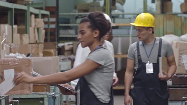 Tracking of female Latin-American factory product manager walking in warehouse inspecting goods and writing something down on clipboard