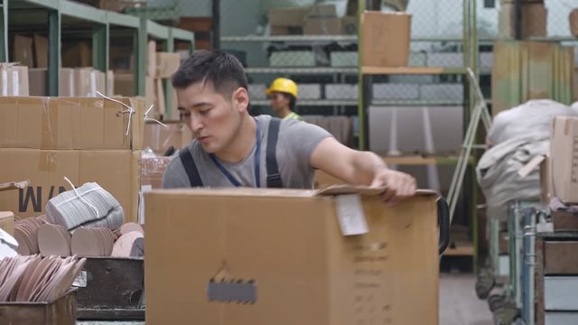 Tired Asian factory worker pushing trolley with cardboard box, then stocking it in warehouse 