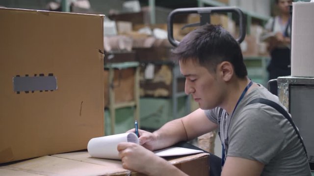 PAN of Asian worker sitting in warehouse and doing paperwork on tip of cardboard boxes