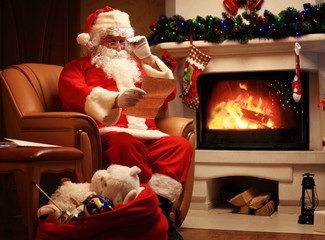 happy Santa Claus sitting at his room at home near Christmas tree and big sack and reading Christmas letter or wish list