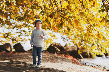 Little boy playing by the lake on a sunny day, wearing warm hat, grey knitted pullover, trousers and shoes