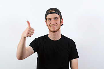 Fototapeta premium Smiling Young Adult Male in Dark T-Shirt and Baseball Hat Worn Backwards Shows His Thumbs Up