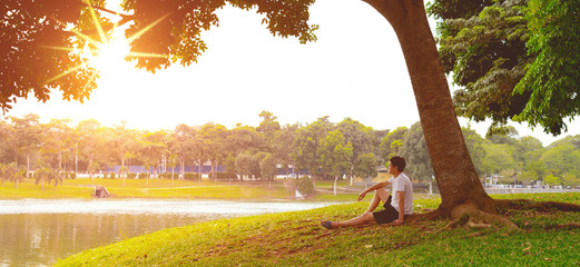 man wearing white t-shirt and black short pants seats on grass under a tree looking at scenic lake view  