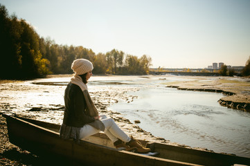 Slim girl in blue jeans, brown jacket and boots, sat on the upturned boat, on the shore, yellow autumn leaves bright sunny day