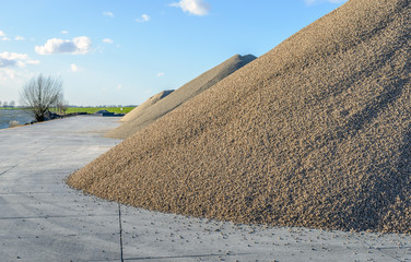 Large heaps of sand and gravel stored on concrete slabs next to a wide Dutch river. It is a sunny day in the middle of the winter season.