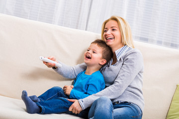 Happy mother and son are sitting on sofa and watching tv together.
