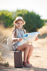 Cute little girl,long hair braided into two pigtails,on his head wearing a sun hat,behind wears light leather backpack,sitting on the old suitcase among the back roads,exploring the map of the area