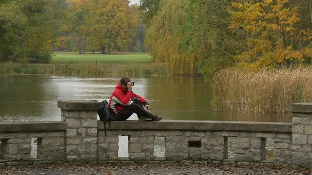 Lonely Tourist in vr Glasses Sittning on Bridge Fence Watching Video 360 Degrees Playing Virtual Games in Cloudy Day at the River or Lake Autumn Park