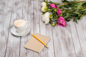 Notebook with a pencil next to coffee and peonies flowers on wooden background.
