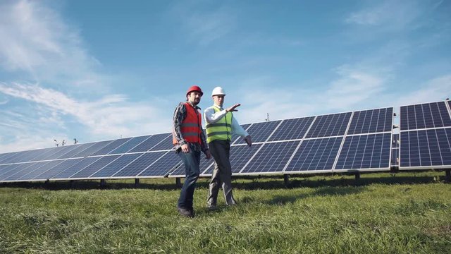 Two male electrician workers in reflective vests and hard hats walking in between long rows of photovoltaic solar panels and talking about installation of new solar panels