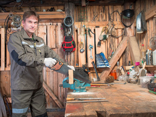 Adult man in work clothes and gloves cost around a wooden table with a vise and processing a piece of wood against the background of the repair shop with tools