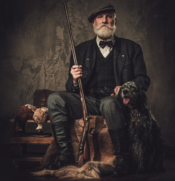 Senior hunter with a english setter and shotgun in a traditional shooting clothing, sitting on a dark background.