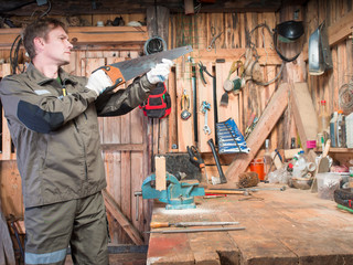 Adult man in work clothes and gloves cost around a wooden table with a vise and checks the blade sharpness against the background of the repair shop with tools