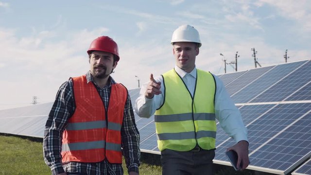 Pair of male maintenance workers in green and white reflective vests standing in front of large solar panels outside