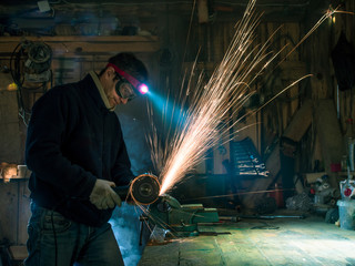 Toned image adult man in protective mask and gloves standing near wooden table with a vise and cutting machine saw off a piece of iron on the background of sparks inside the repair shop with tools - obrazy, fototapety, plakaty