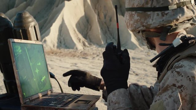 Soldier is Using Laptop Computer for Tracking the Target and Radio for Communication During Military Operation in the Desert