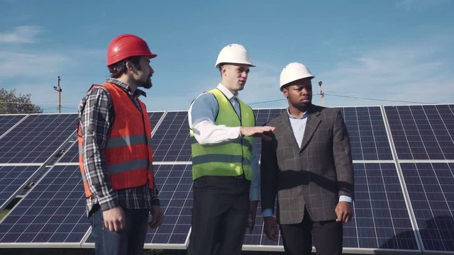 Solar panel workers and manager in red and white hard hats standing and talking and handshaking outside with large power arrays behind them