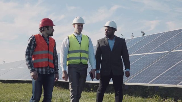Two male electrician workers in reflective vests and hardhat and one investor walking in between long rows of photovoltaic solar panels