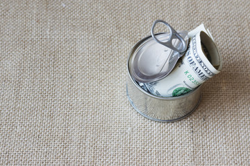 Dollar Bills rolled up inside opened round tin with copy space. Subjects captured against soft window lighting on linen fabric  background.
