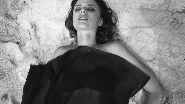 Sensual blonde woman lying at the sandy beach while black see through fabric covers her body from bottom to the top of her head - black and white video in slow motion