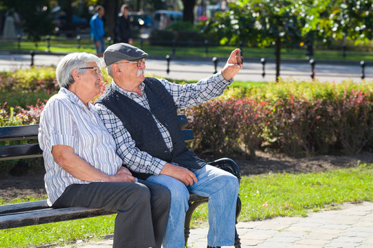 senior couple taking selfie outdoors, happy mature pair having fun while sitting on a bench in autumn day in a park