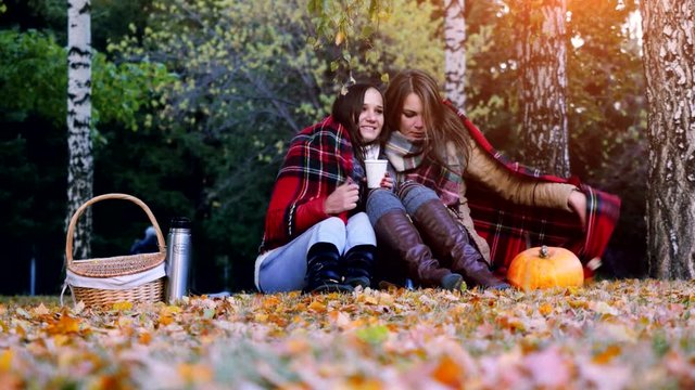 Young beautiful women sitting on picnic bundled up in a blanket drinking hot tea from thermos in autumn park. Girls rug near the pumpkin of Halloween theme. 3840x2160