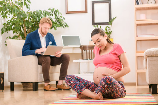 Picture of pregnant woman sitting on floor and touching her belly during training. Brunette lady doing yoga while her husband businessman busy working on laptop computer.